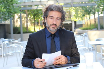 Mature man daydreaming outside the office sitting on outdoors table  with a document in his hands staring up into space lost in thought