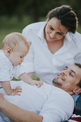 Happy young family having fun, sit and lay on the grass, bowl, rising up, piggyback ride their child in park on summer sunset
