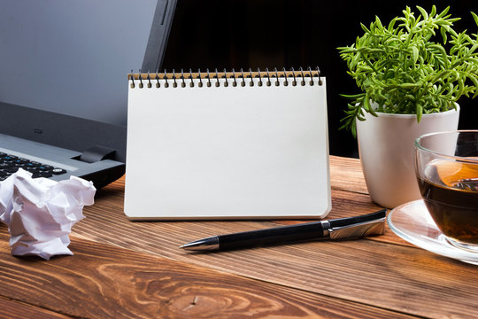 Office table desk with supplies, white blank note pad, cup, pen, pc, crumpled paper, flower on wooden background. Top view