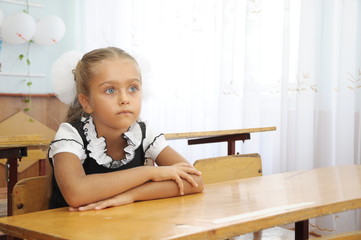 September 1 schoolgirl at the desk. Beautiful girl first grader in school uniform sitting at a desk in classroom. Schoolgirl with folded hands sitting at school desk at lesson, copyspace. 

