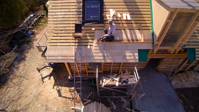 A roofer man installing shingles on the other part of the roof of a wooden cabin house with trees on the side