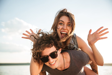 Young happy smiling couple in love piggyback at the beach - obrazy, fototapety, plakaty