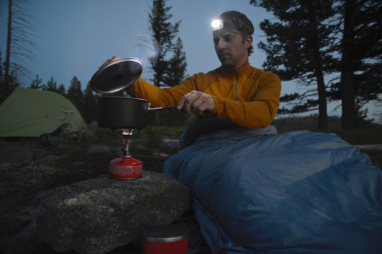 Male camper in sleeping bag prepares meal on camping stove at night on Midnight Ridge, Colville National Forest, Washington State, USA