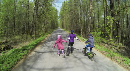 Fototapeta premium Mother with children ride on rollerblades and bike by road with bicyclists at sunny spring day in forest. Aerial view