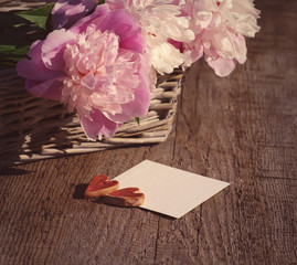 Two hearts on the white empty card for an inscription and a bouquet of pink peonies on a wooden surface, close up