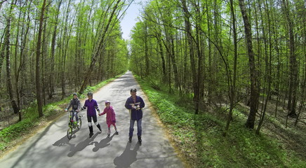 Four people parents and children ride on rollerblades and bicycle by road along forest at sunny spring day. Aerial view