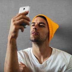 Attractive, handsome and serious young man , reading someting, taking selfie. Wearing orange hat and gray scraf on gray background.