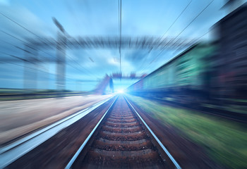 Naklejka premium Railway station with cargo wagons and train light in motion at sunset. Railroad with motion blur effect. Railway platform. Heavy industry. Conceptual background