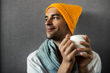 Dreaming, smiling and sitting young handsome man with a mug of coffee, tea, water, on gray background. Wearing orange hat and gray scarf.