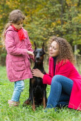 Naklejka na ściany i meble Little curly girl and her mother are caress the dobermann in park.