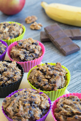 Fruit muffins on a yellow napkin and a grey wooden table. Apple, banana, walnuts and chocolate in the background.