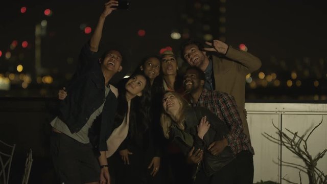 Happy mixed ethnicity friends pose for a selfie on city rooftop at night
