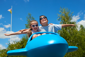 child girl and boy fly on blue plane attraction in city park, happy childhood, summer vacation...