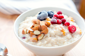 Oatmeal porridge with dried apricots, blueberries, cranberries and chopped almonds on bright wooden table. Jar of honey on background. Selective focus. Bright healthy breakfast image