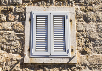 Closed PVC window with shutters on the wall of the old building of white coquina (shell limestone) in Old Town of Budva, Montenegro. Connection of modern building technologies with antiquity