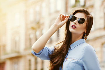 Outdoor portrait of a young beautiful confident woman posing on the street. Model wearing stylish sunglasses. Girl looking up. Female fashion. Sunny day. Close up. City lifestyle. Copy space for text 