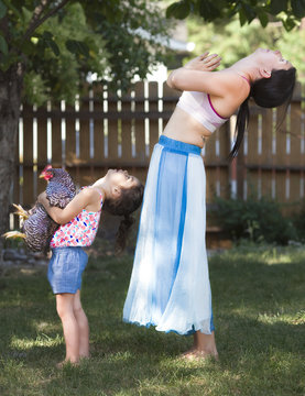 Mother And Daughter Practicing Yoga With Chicken In Backyard