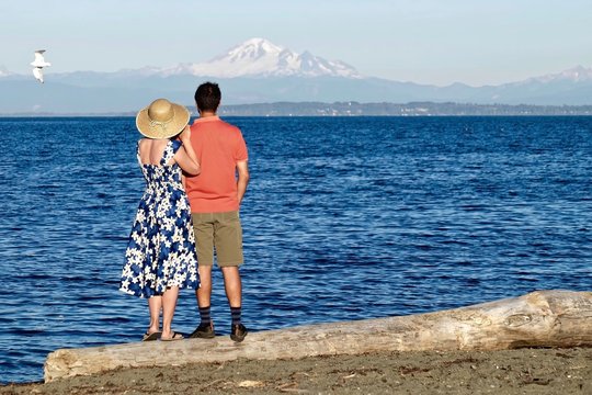 Married couple on beach huggin and looking at view of snow capped volcano. Centennial Beach in Boundary Bay Regional Park, Tsawwassen, BC, Canada. 
