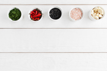 Food seasoning variety on white wooden background, flat lay. Bowls with black and pink sea salt, parsley, chili and garlic, free space. Natural organic flavoring, cooking concept