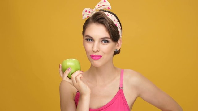 Beautiful cute smiling pinup girl in yellow dress eating green apple and showing thumbs up gesture