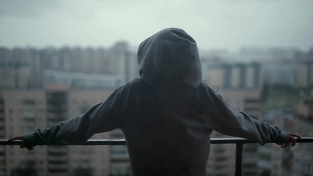 Girl in a hoodie on the balcony and looking at the high-rise buildings. It's raining.