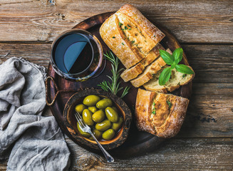 Wine snack set. Glass of red wine, green mediterranean olives, freshly baked ciabatta bread in dark wooden plate over rustic wooden background. Top view, horizontal composition