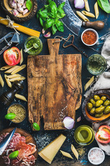 Italian food cooking ingredients on dark background with rustic wooden chopping board in center,...