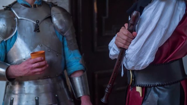 A knight is holding a pottery cup in his hand and a member of the royal family is holding a flute. They are talking to each other. Close-up shot.

