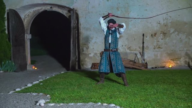 A young man is swinging his whip as a part of the performance. He is also having one hand on his ear. Wide-angle shot. It is evening time.
