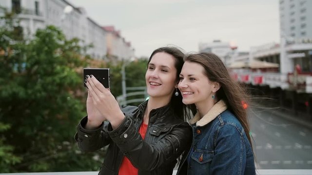Two girls best friends taking selfie, standing on the city bridge, talking, smiling, laughing. slow mo.