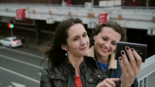 Two girls best friends taking selfie, standing on the city bridge, talking, smiling, laughing. slow mo.