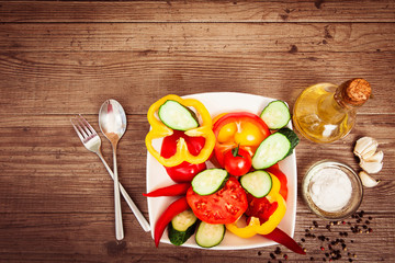 fresh vegetables salad with tomatoes, cucumbers, sweet peppers and chilli in a deep white plate. Seasoning, olive oil, garlic on wooden table background. Copy space above. 