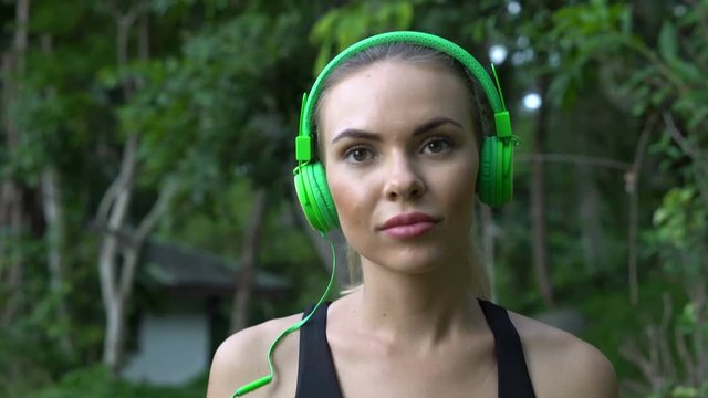 Close uo of young pretty blonde fitness female wearing bright green headphones, listening music before run walking on the beach and enjoying beautiful summer day