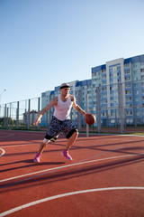 Fototapeta premium Guy plays basketball on a red Court in sunny weather the background of houses.