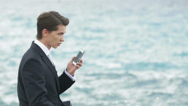 Handsome businessman in suit standing on the rocky shore and talking by phone. Sea waves on background