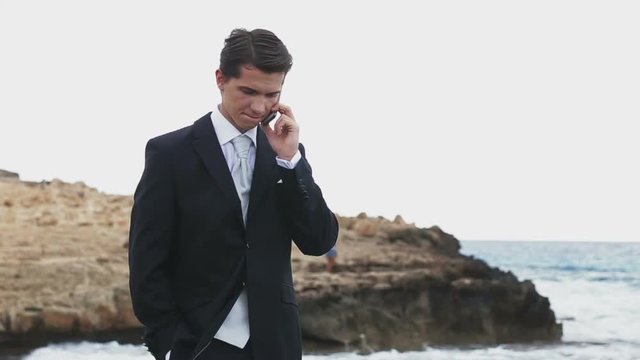 Handsome businessman in suit standing on the rocky shore and talking by phone. Sea waves on background
