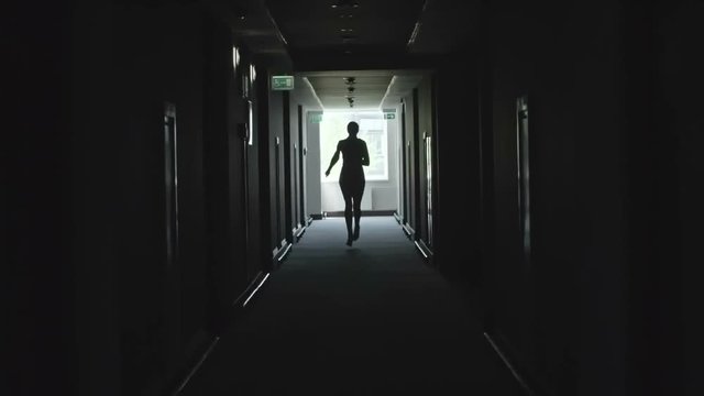 Black silhouette of businesswoman running alone through dark corridor in office building and holding documents in slow motion