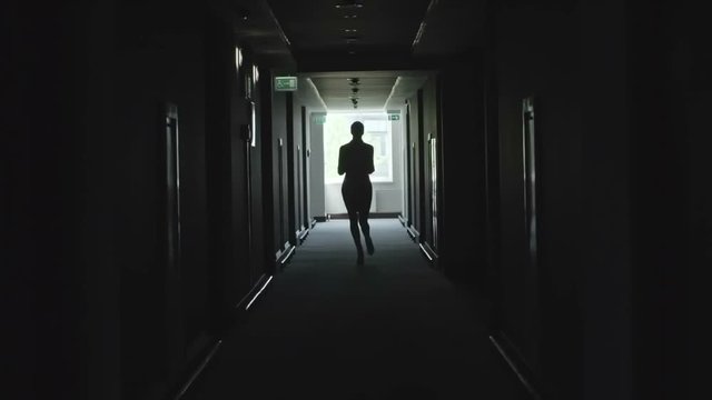 Silhouette of businesswoman with documents dropping them on the floor while running through dark corridor in office building 