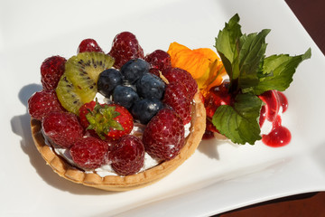 Tartlet with cheese cream, wild berries and kiwi, on a white plate, decorated with orange flower, leafs, cream and jam, served on a wooden table