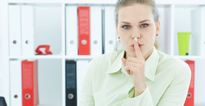 Portrait of an attractive business woman with finger on lips. Young businesswoman in office asking for silence. Woman with finger on lips gesturing for quite.