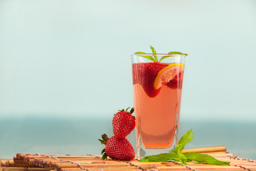 Glass of strawberry lemonade with pieces of strawberry, lemon and fresh mint. Closeup of mojito with beach on background. Fresh summer bright strawberry cocktail.