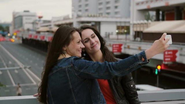 Two girls best friends taking selfie, standing on the city bridge, talking, smiling, laughing. 4K