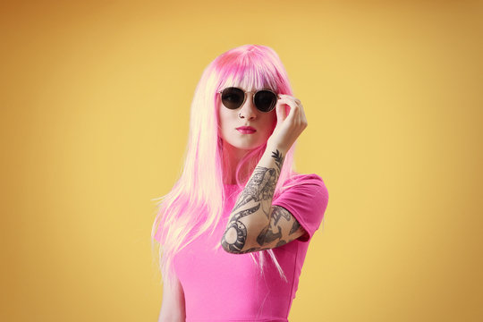 Beautiful young woman with tattoo wearing pink wig and sunglasses on yellow background
