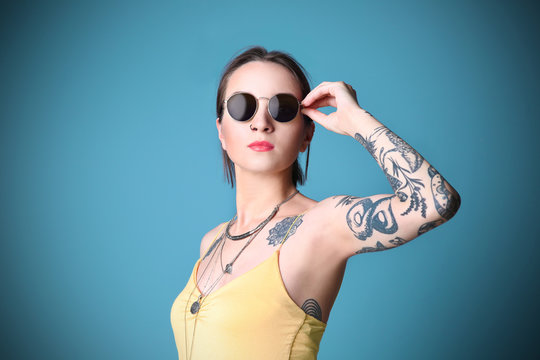 Beautiful young woman with tattoo wearing sunglasses and posing on blue background