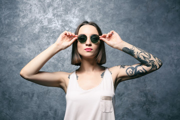 Beautiful young woman with tattoo wearing sunglasses and posing on gray background