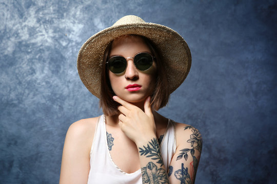 Beautiful young woman with tattoo wearing sunglasses and hat on gray background