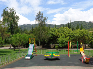 City playground, small slide, swings and carousel on green lawn with trees