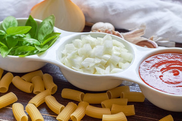 Basil, sliced onions and tomato paste in white bowl with raw pasta and garlic scattered around on wooden background. Concept image for Italian cuisine