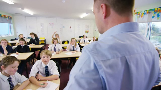  Young students listening to teacher & putting hands up in school classroom