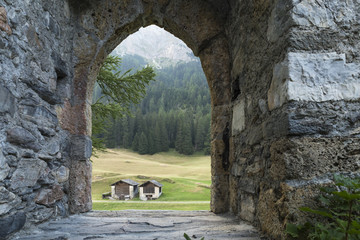 Blick durch den leeren Torbogen einer alten Burgruine auf zwei Strohspeicher. Eine grüne Wiese und...